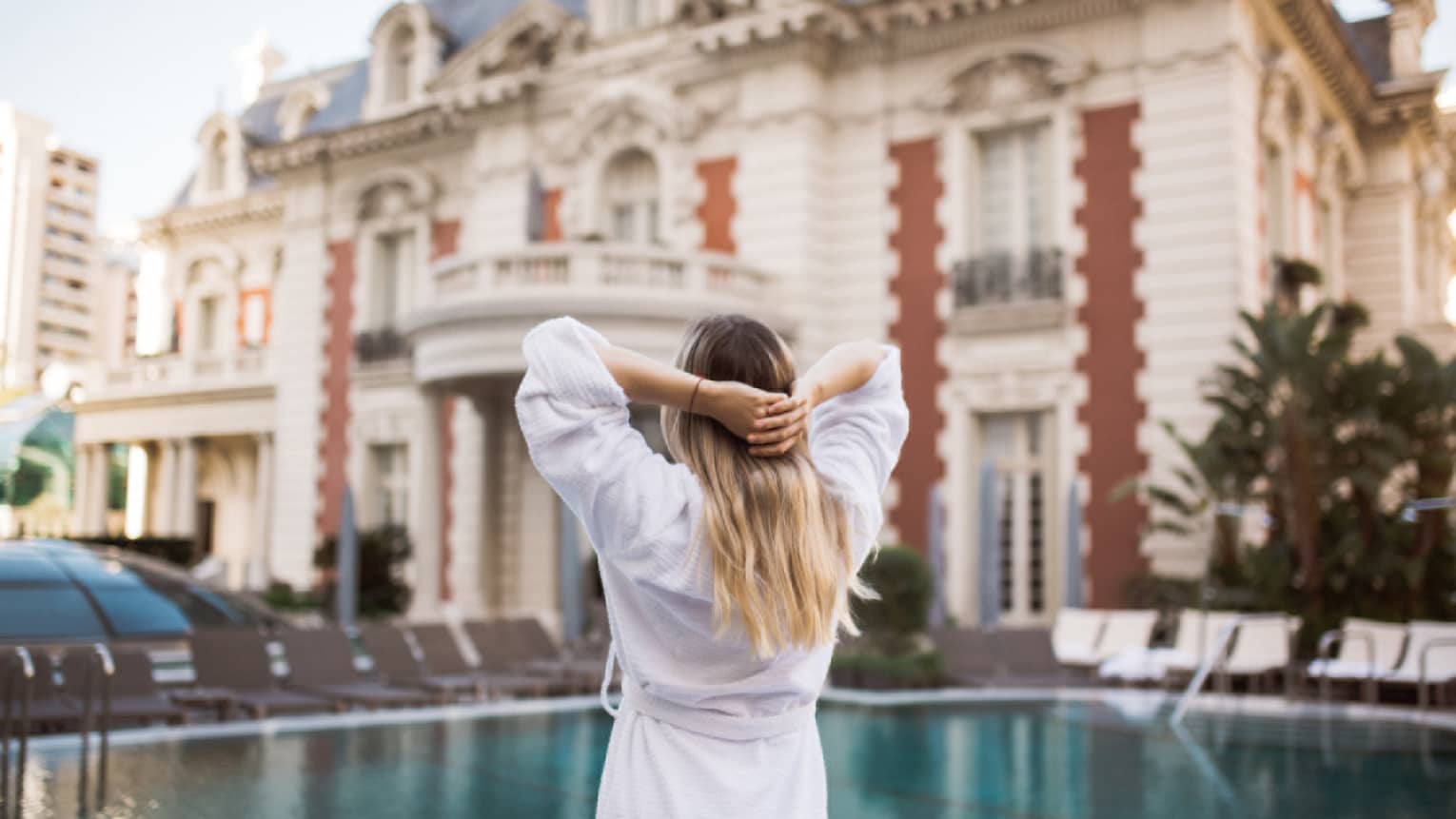 Back of woman with hands behind head by outdoor swimming pool and mansion exterior