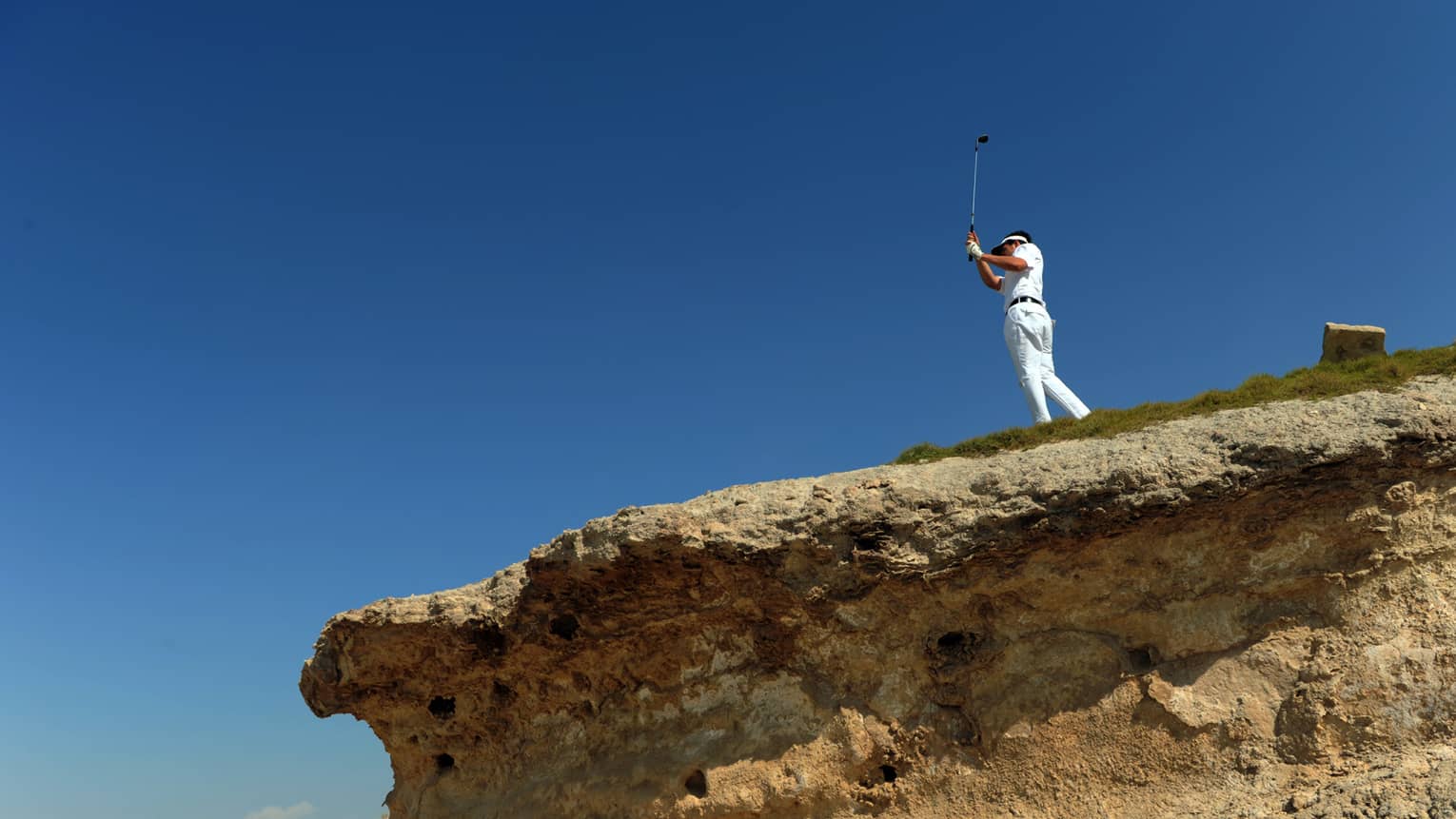 Man swinging golf club on top of rock cliff against blue sky