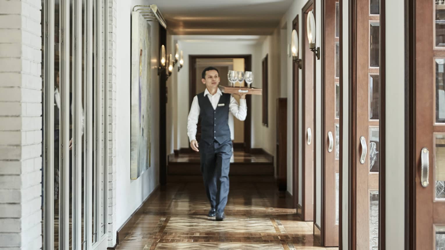 Hotel staff walks down hall, carries tray with empty wine glasses 