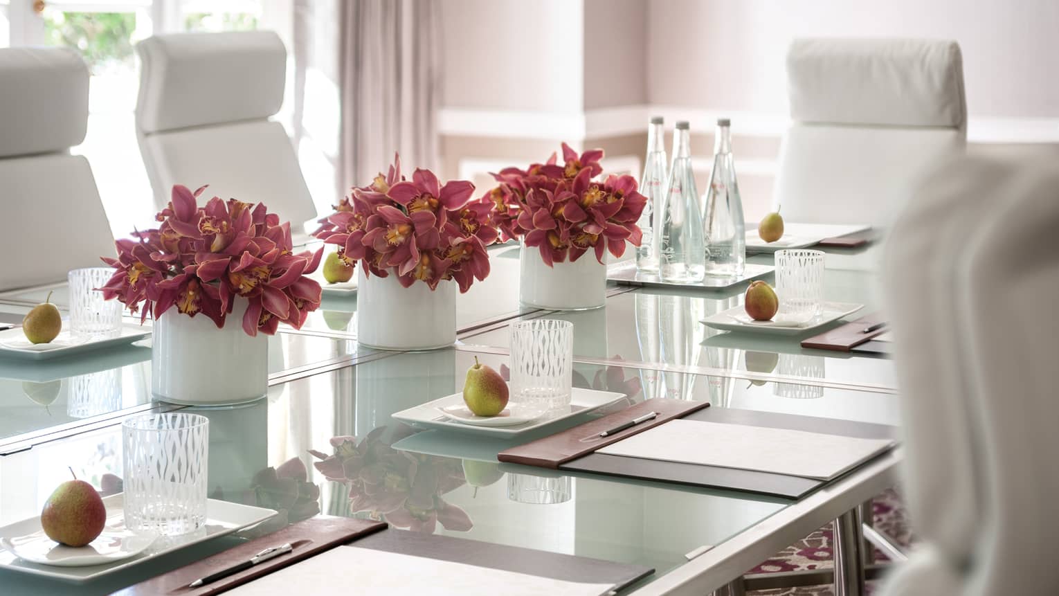 Glass meeting table set with pink flowers in vases, fresh pears and agendas in front of white leather chairs 