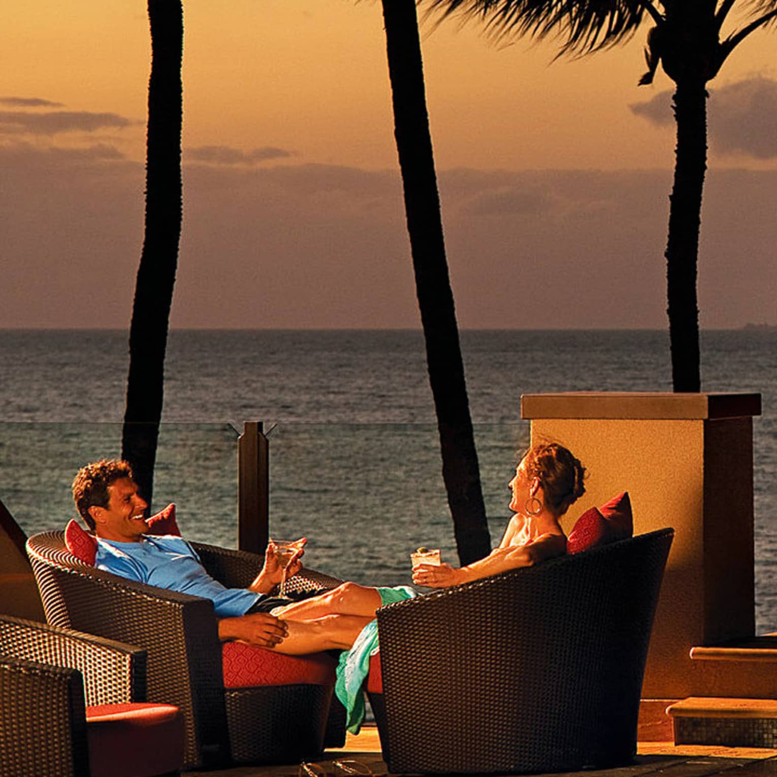 Man and woman sit in wicker patio chairs with cocktails at sunset, next to infinity pool and fire pit 