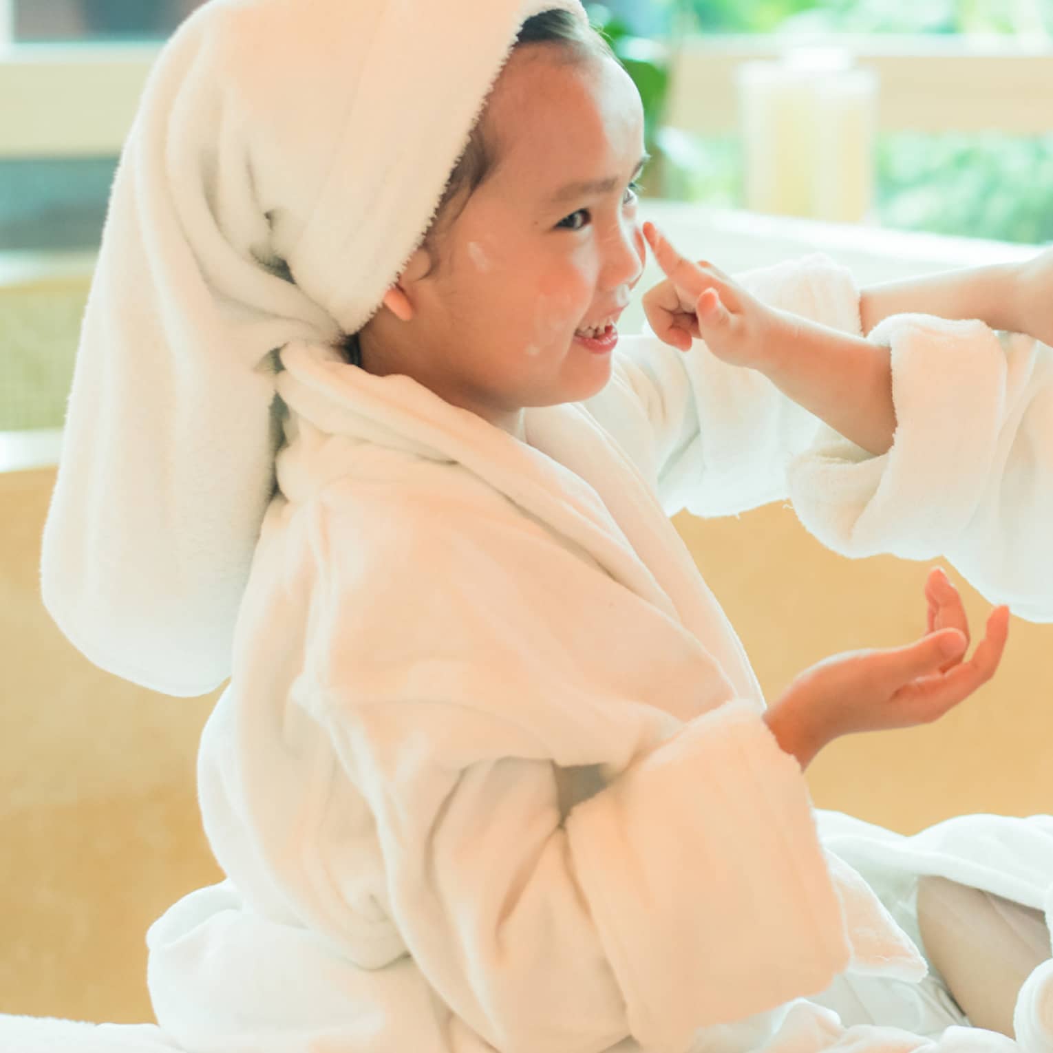 Two laughing young children in white bathrobes with towels around hair put lotion on each other's faces 