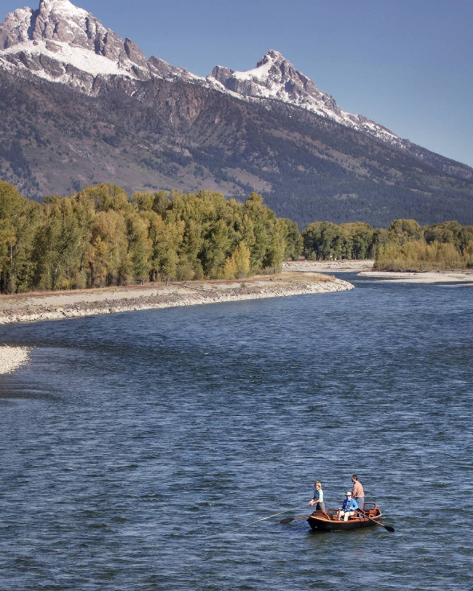 People fishing in boat on wide river by rocky shore, green trees, mountains