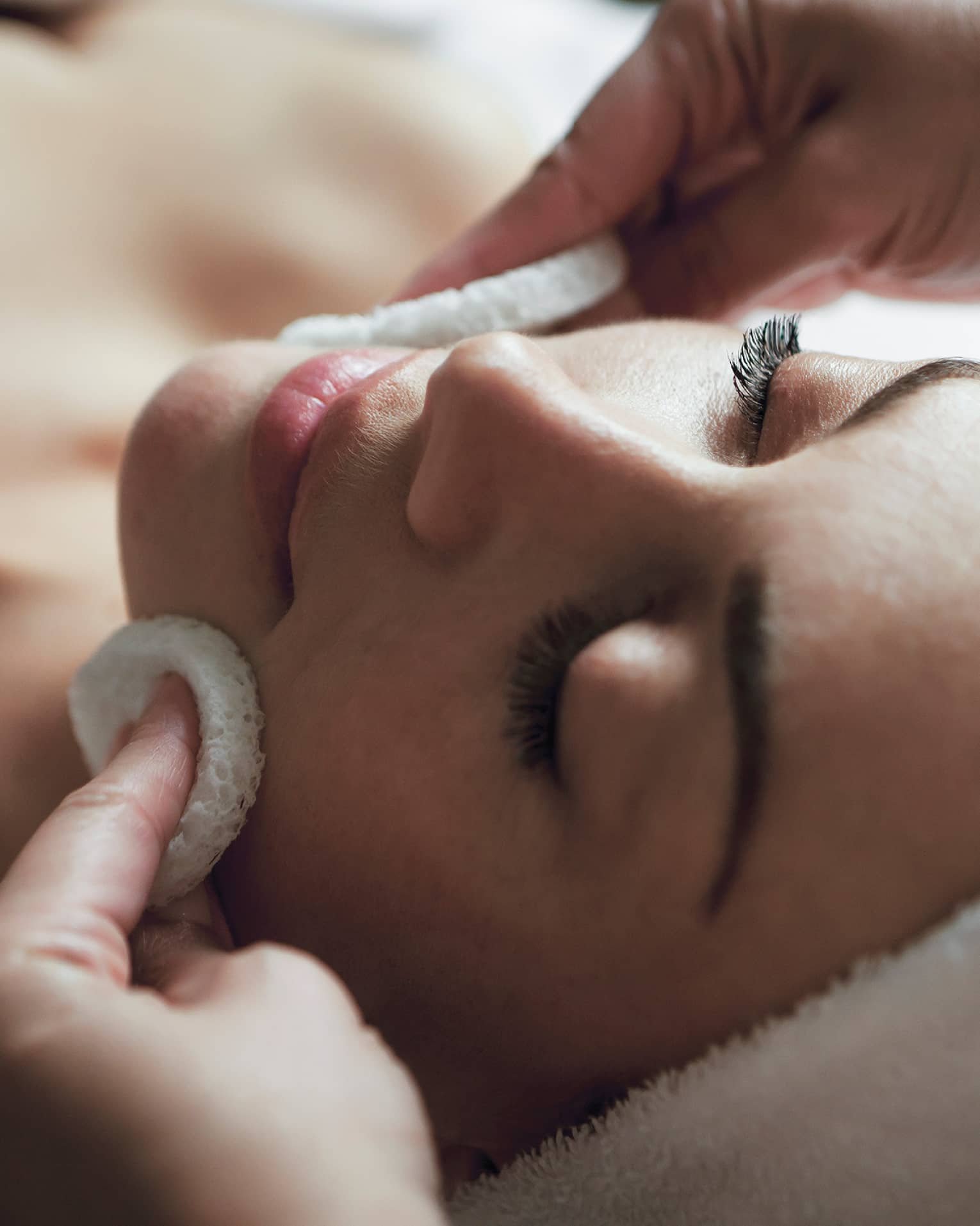 Spa staff applies facial cleanser to woman's face, her eyes closed and towel around hair