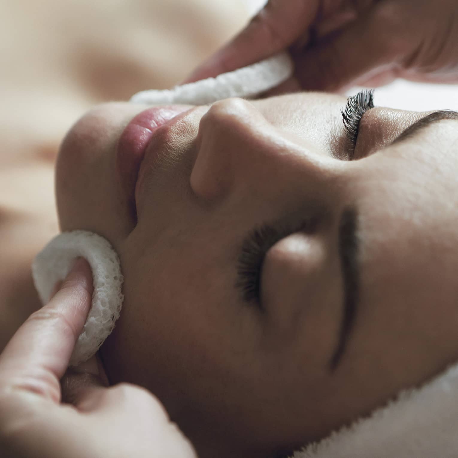 Spa staff applies facial cleanser to woman's face, her eyes closed and towel around hair 
