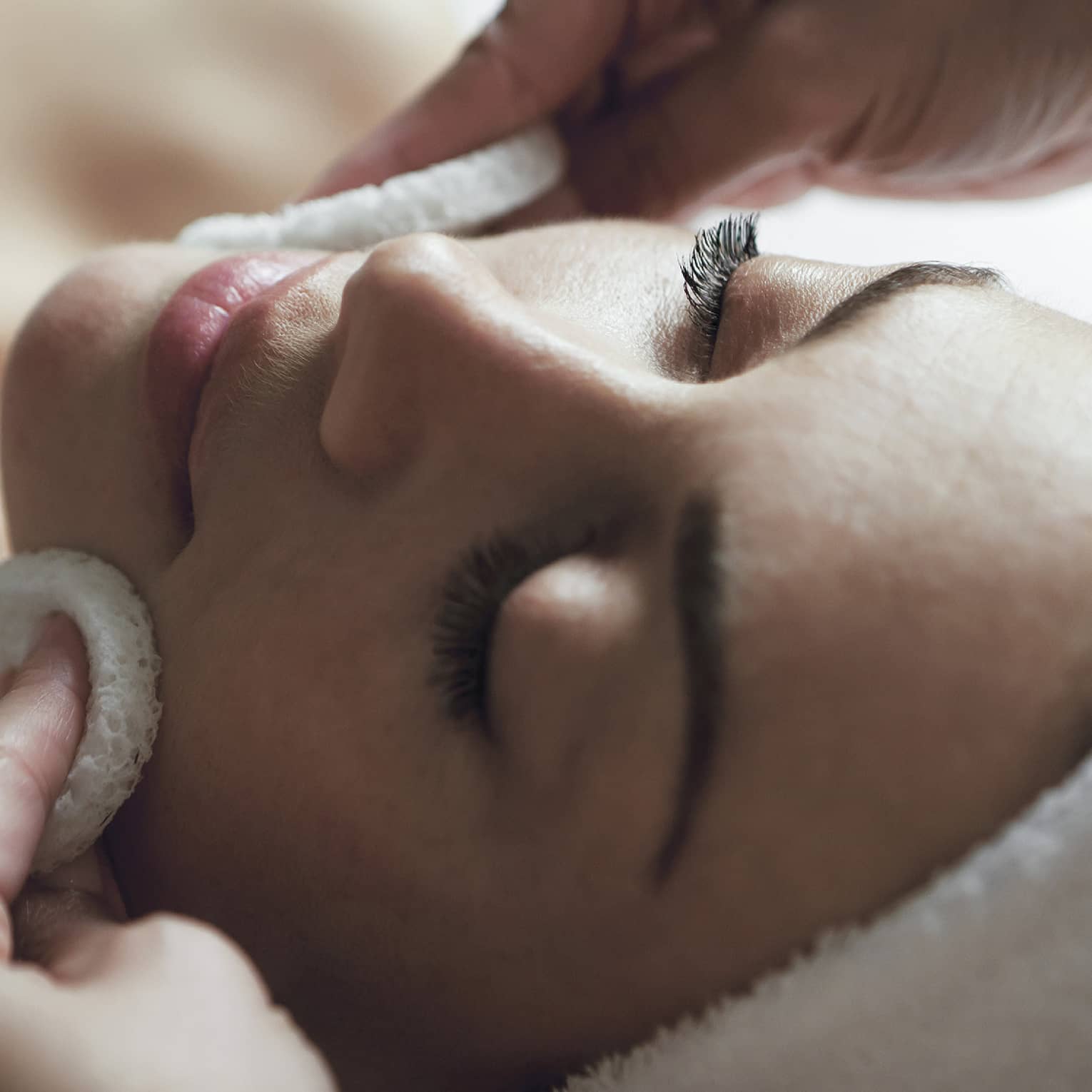 Spa staff applies facial cleanser to woman's face, her eyes closed and towel around hair 