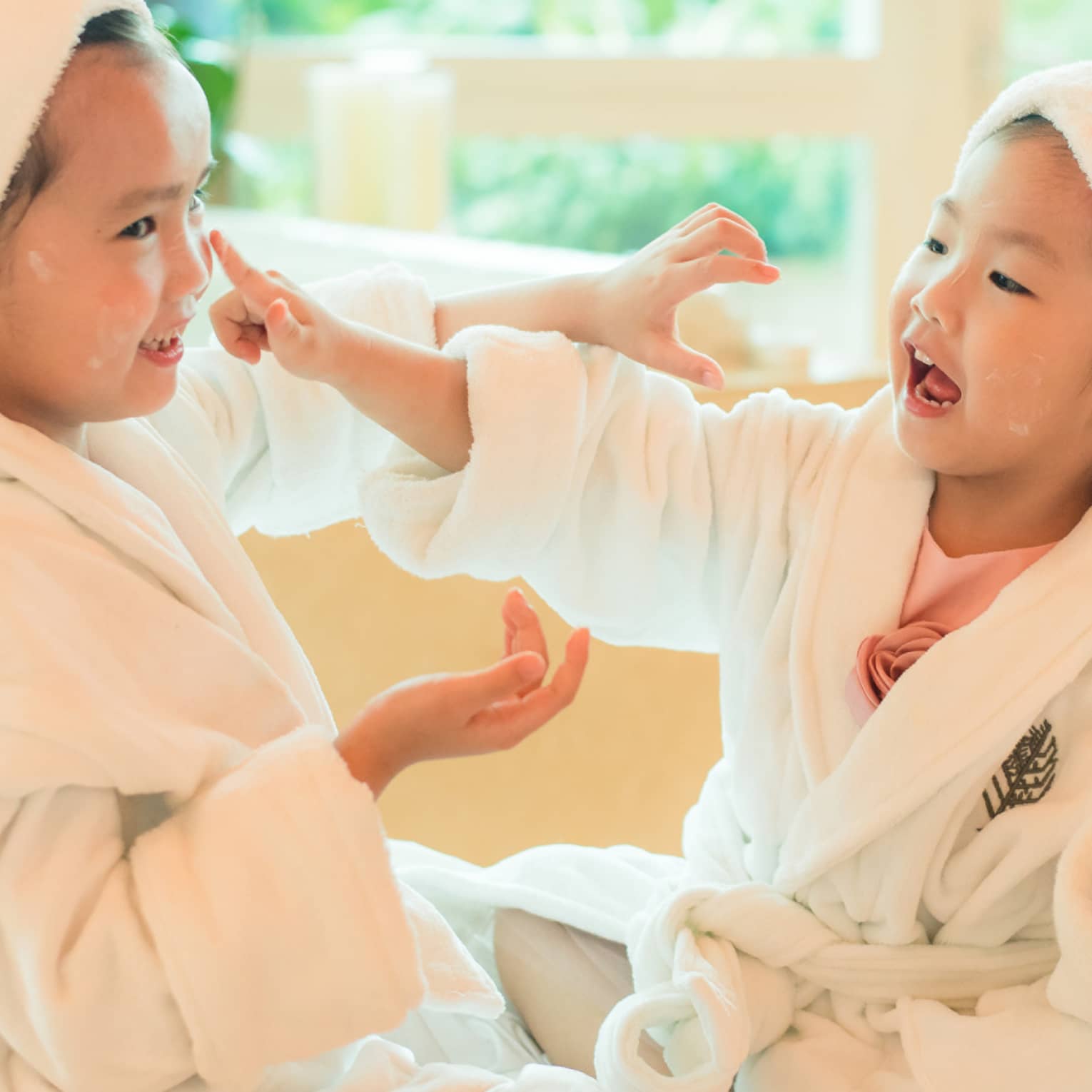 Two laughing young children in white bathrobes with towels around hair put lotion on each other's faces 