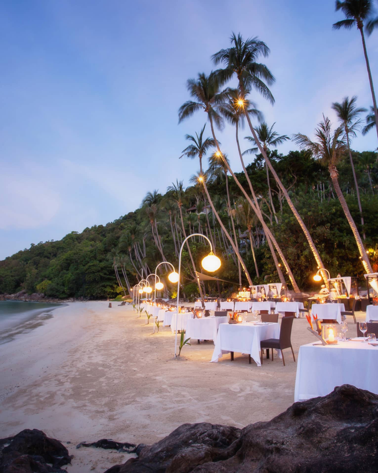 Dining tables set up along white sand beach under palm trees at dusk