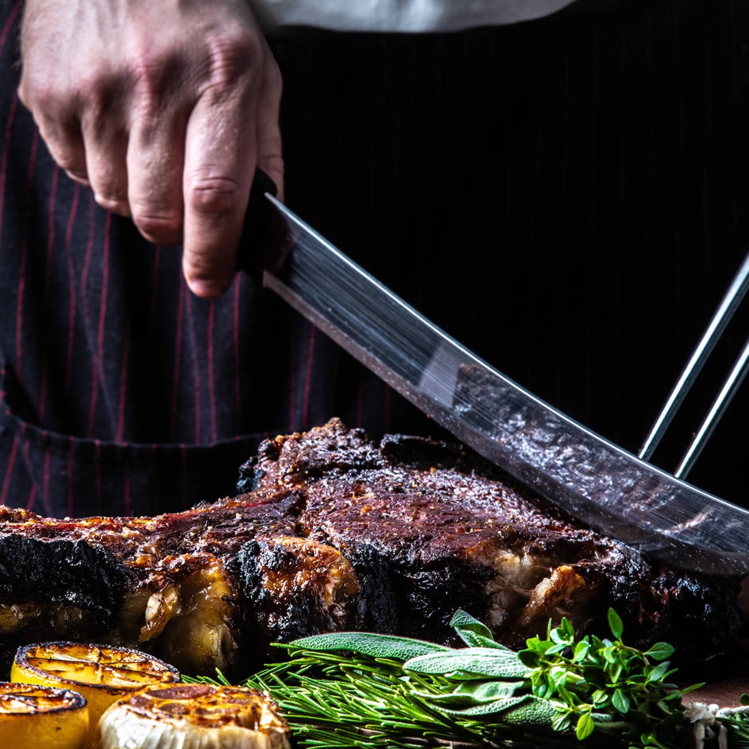 Close-up of chef slicing large rack of grilled meat, vegetables with butcher knives