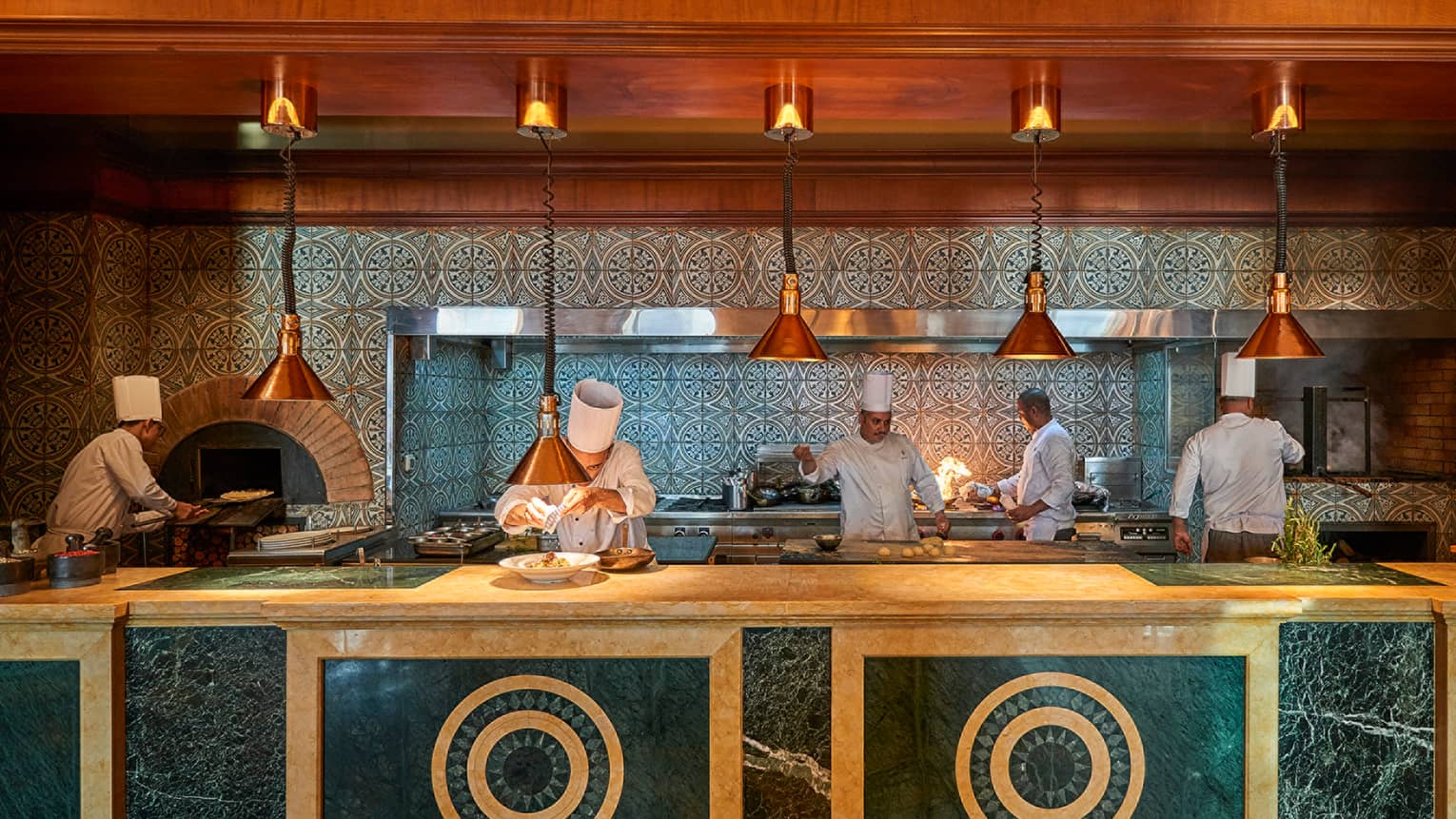 Bella chefs in white coats and hats prepare meals on large cream-and-brown marble counter under brass lamps