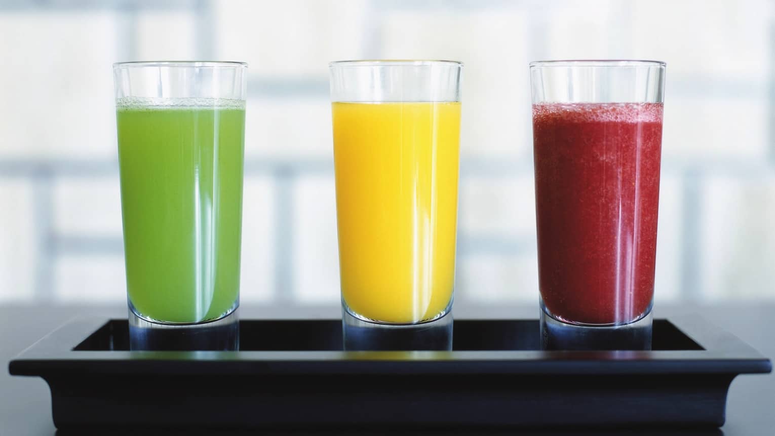 Three tall juice glasses with colourful green, orange and red smoothie drinks on tray