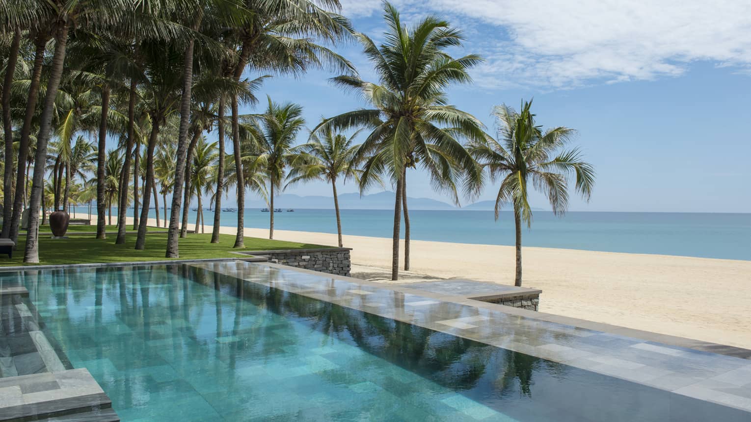 Tall palm trees between stone outdoor swimming pool and white sand beach