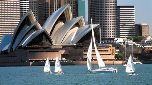 Four sailboats float in harbour in front of Sydney Opera House with modern, angular roof