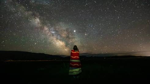 A lone woman stargazes, wrapped in a colourful striped blanket 