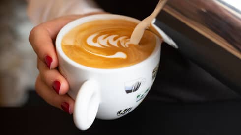 Close-up of hand holding and stirring white mug with foamy latte coffee