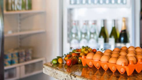 Smiling man holds green bottle in front of open fridge, fresh eggs on counter