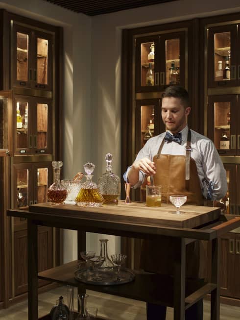 Bayou & Bottle mixologist in apron stirs bourbon cocktail, surrounded by illuminated display cases with bottles 