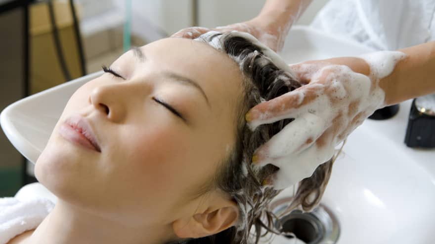 Woman closes eye and leans back against sink as hands massage shampoo into her scalp