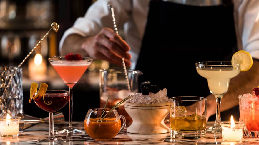 Mixologist stirs cocktails on bar top with various colourful drinks, wood bowl with crushed ice