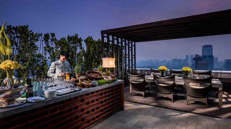 Bartender prepares drinks, cheeses, salads at long Imperial Suite rooftop bar by private dining table at night