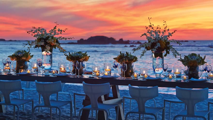 Long candle-lit wedding table on white sand beach under vibrant sunset