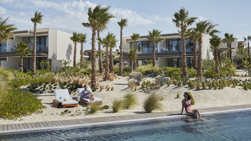 A woman sits on the edge of a pool and looks back at her partner sitting in a lounge chair just beyond the hotel