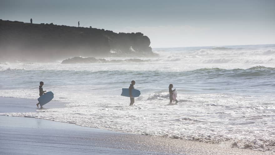 Three people carry surfboards into tide on Portugal beach