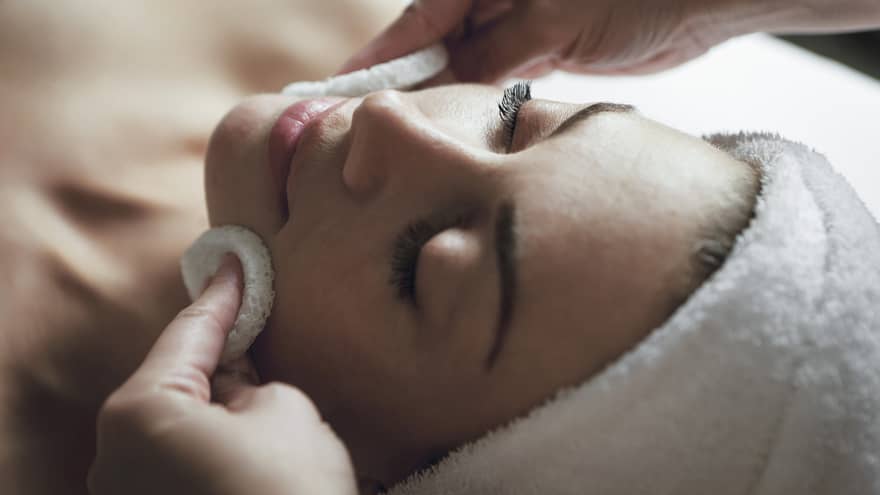 Spa staff applies facial cleanser to woman's face, her eyes closed and towel around hair 
