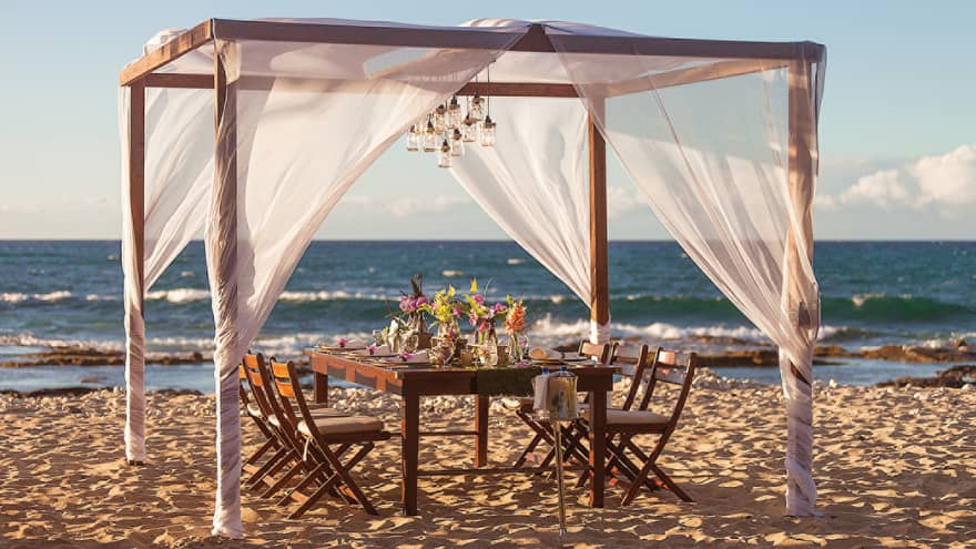 Sheer white linens on wood posts over private dining table on sand beach