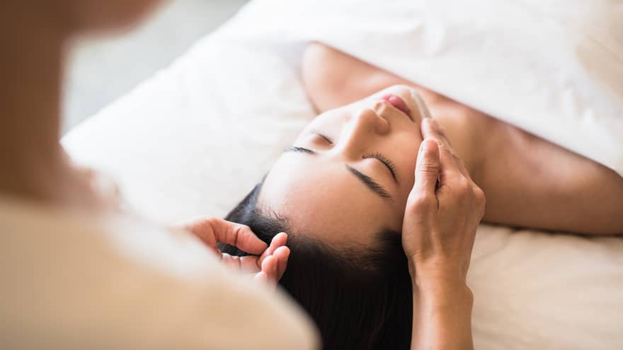 Woman lying under white sheet on massage table, eyes closed as spa staff rubs lotion on her chin