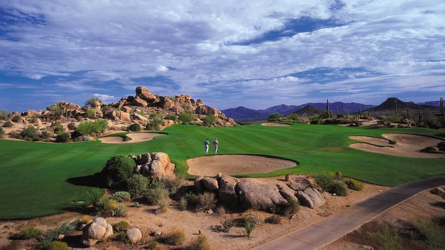 Two men on vast, green desert golf course under sweeping blue sky 