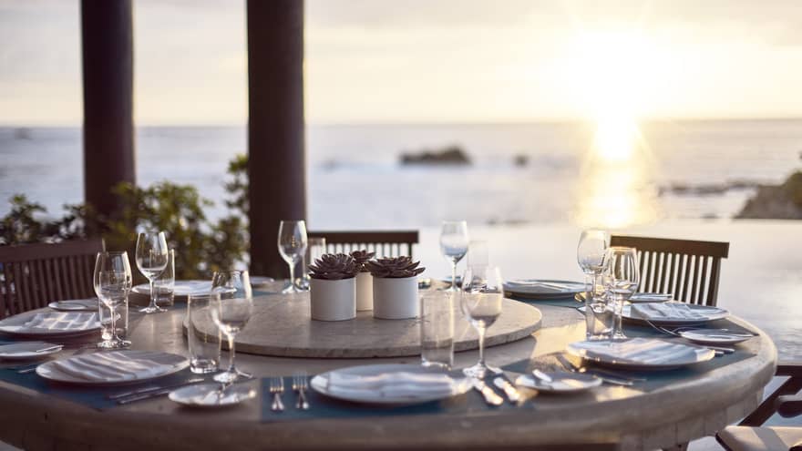Round outdoor dining table with wine glasses, ocean and sunset in background